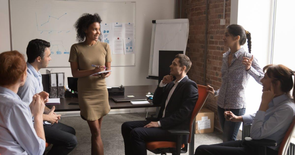 A woman standing and leading a group discussion with seated employees in an office environment during an on-the-job training session.