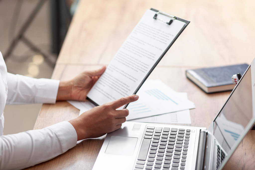 A person reviewing training documents on a clipboard beside a laptop on an office desk.