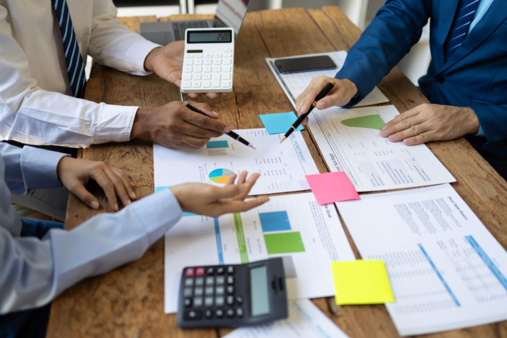 Two colleagues pointing at financial charts, figures, and sticky notes on a desk during a budget review meeting.