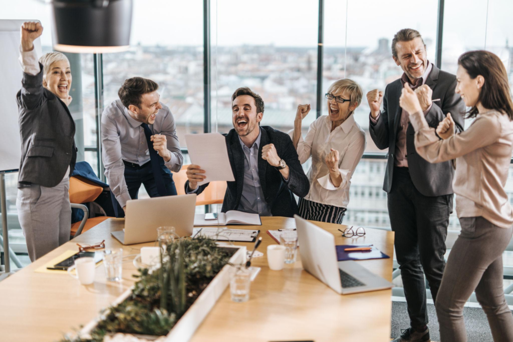 A group of excited business professionals celebrating success and cheering with raised arms around a conference table in a modern office.