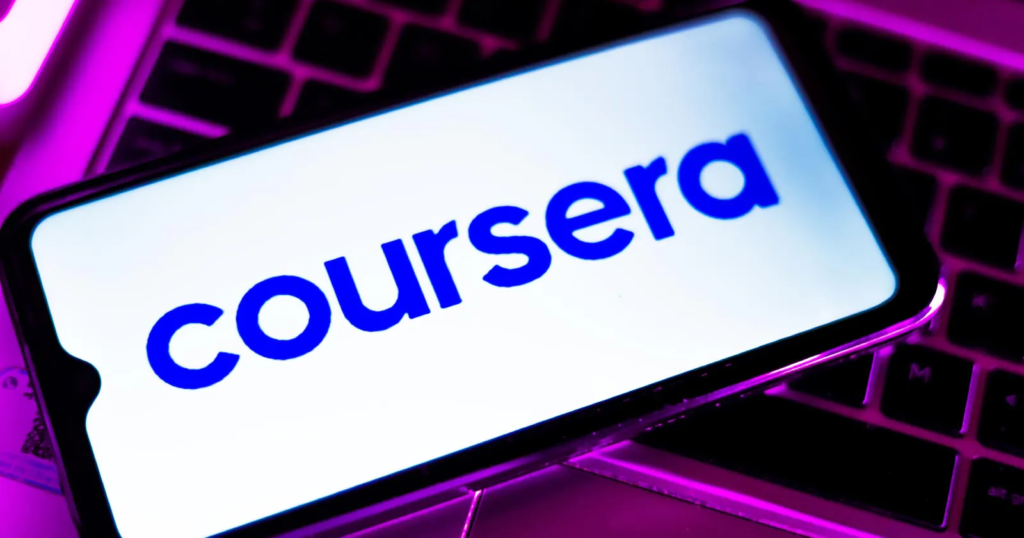 Sebuah telefon pintar memaparkan logo Coursera berwarna biru di atas paparan putih, diletakkan di atas papan kekunci komputer pekena lampu neon merah jambu.